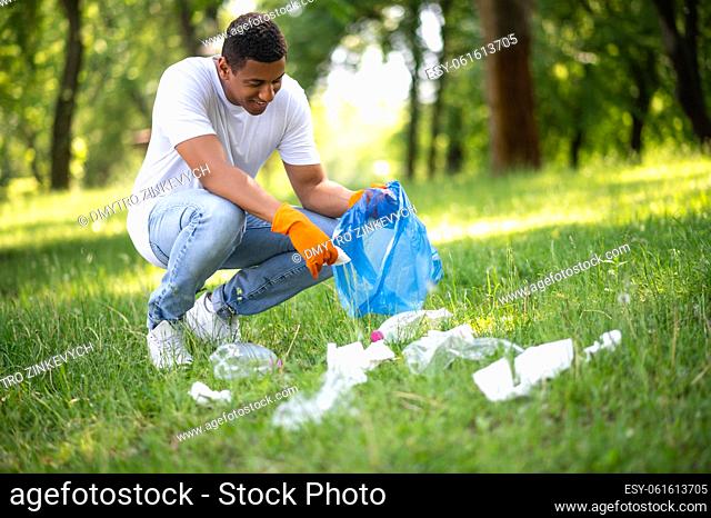 Cleaning, nature. Young adult dark-skinned man in gloves crouched bagging garbage on green lawn on warm day