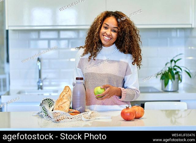 Smiling young woman holding fruit by groceries shopping bag in kitchen