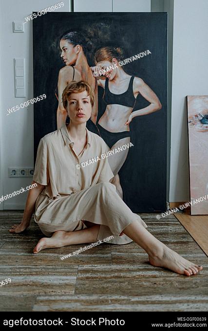Confident mid adult female artist sitting on hardwood floor against painting