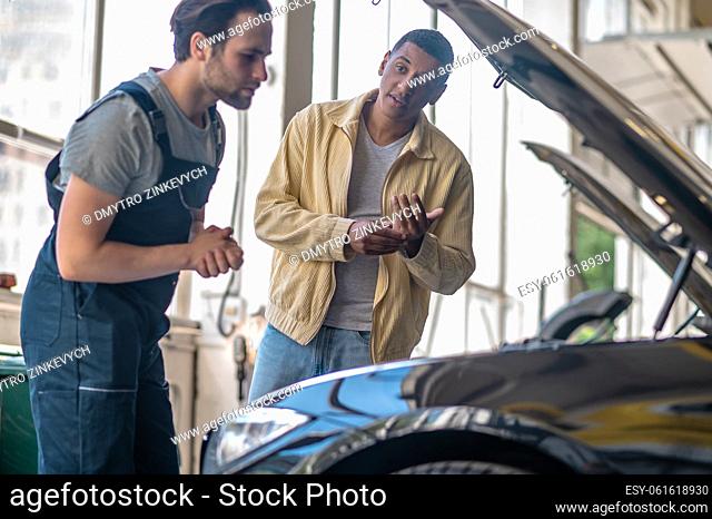 Car service. African american looking questioningly at caucasian serious mechanic looking into open car hood standing in workshop