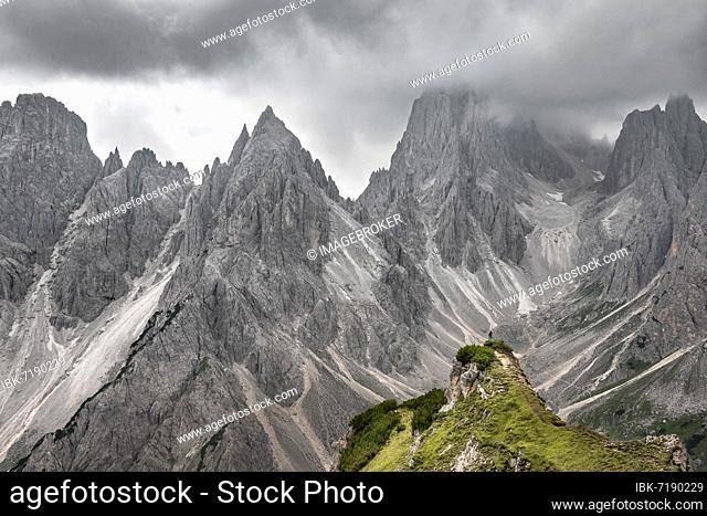 Hiker standing on a ridge, mountain peaks and pointed rocks behind, dramatic cloudy sky, Cimon di Croda Liscia and Cadini group, Auronzo di Cadore, Belluno