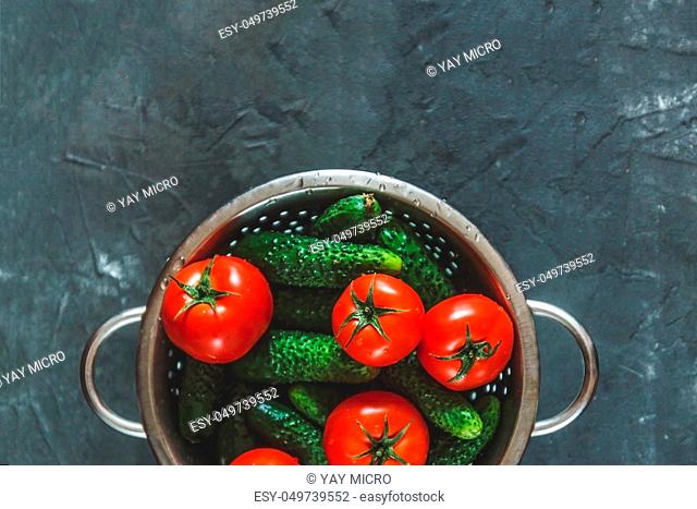 raw whole washed tomatoes and cucumbers in a colander on a dark concrete background. View from above. Flat lay