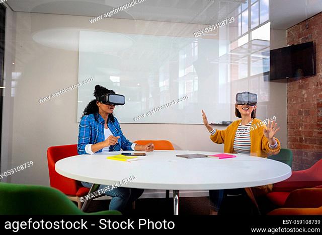 Diverse female colleagues sitting at work wearing virtual reality set and playing