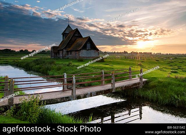 View of grazing moor with footbridge over flooded ditch and church at sunset, St Thomas a Becket Church, Fairfield, Walland Marsh, Romney Marsh, Kent, England
