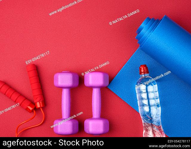 pair of purple plastic dumbbells, bottle of water on a red neoprene mat, top view, training equipment