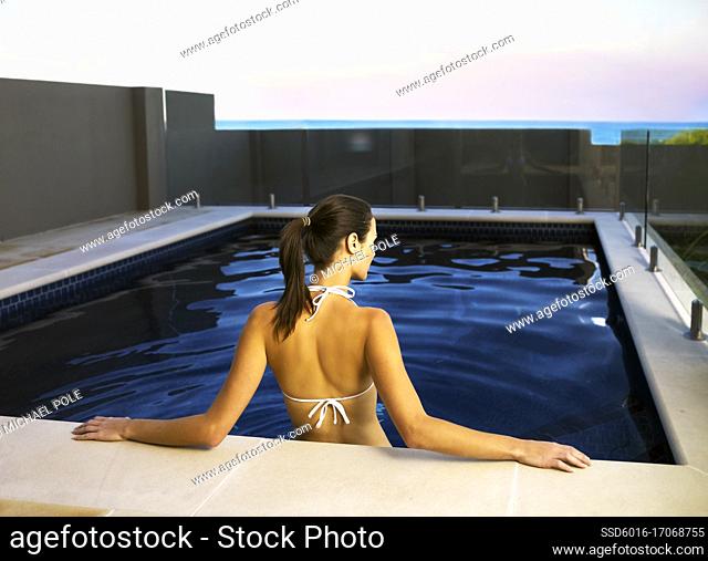 Back view of young woman standing in swimming pool and leaning against the edge with the sea in view over wall