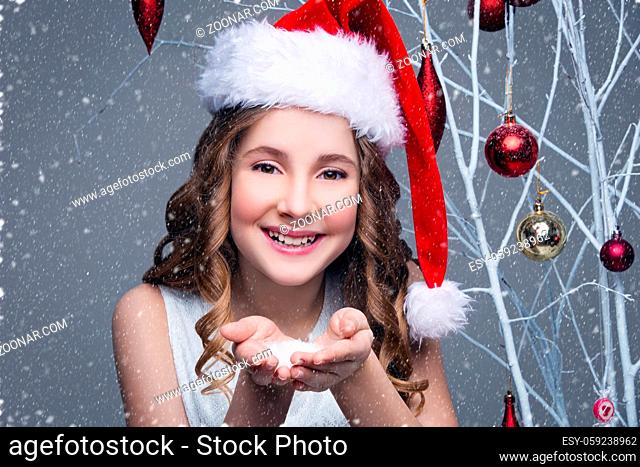Beauiful girl in red santa cap showing snow on palms of hands. Happy expression. Studio shot on christmas ornament background with falling snow