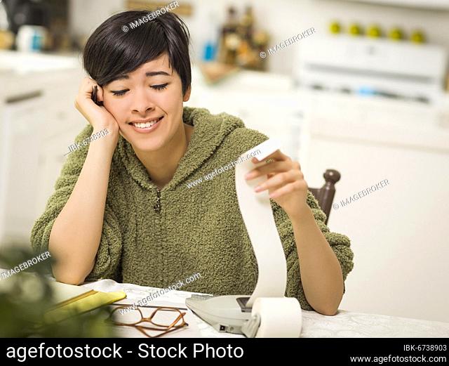 Multi-ethnic young woman relieved and smiling over financial calculations in her kitchen
