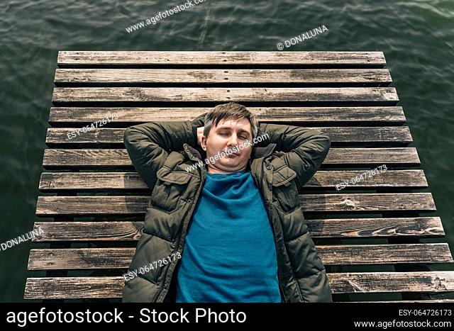 A man lies on a wooden plank pier on the lake in a jacket and jumper with his eyes closed