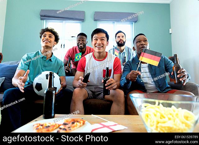 Multiracial curious male friends with beer bottles, flags and ball watching soccer match at home