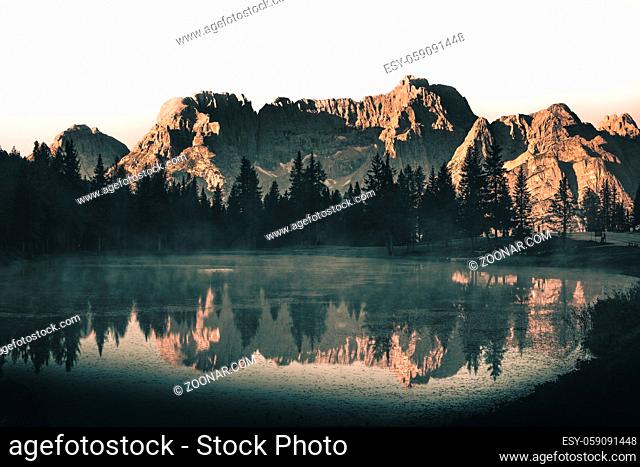 Mountains and sky reflection in a lake with forest in the dolomites during sunrise with fog on the water