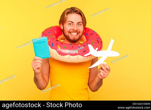 Excited optimistic funny tourist guy in undershirt standing with pink donut rubber ring, holding passport document and airplane mockup, rejoicing travel tour