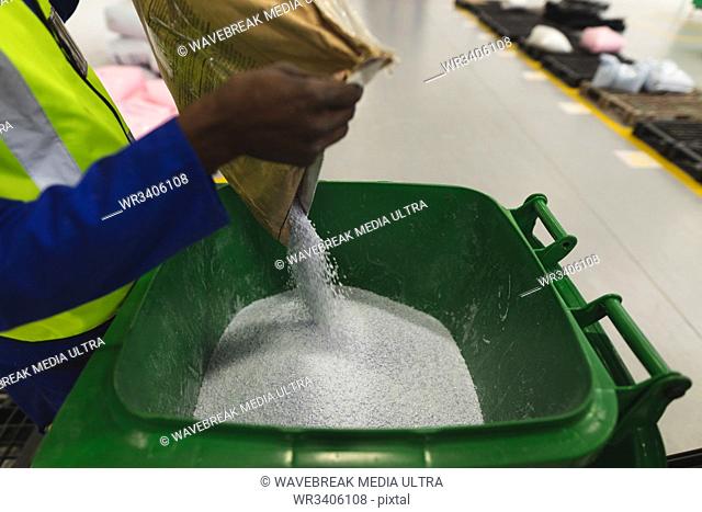 Side view mid section of an African American male factory worker pouring a white granules from a bag into a green plastic bin container to a bucket in a...