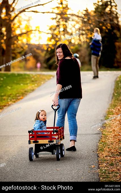 A mother pulling her baby with Down Syndrome in a wagon along a river in a city park with her family during the fall season; St