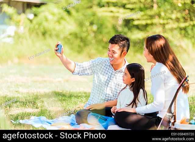 Happy Asian young family father, mother and child little girl having fun and enjoying outdoor sitting on picnic blanket taking selfie using technology mobile...