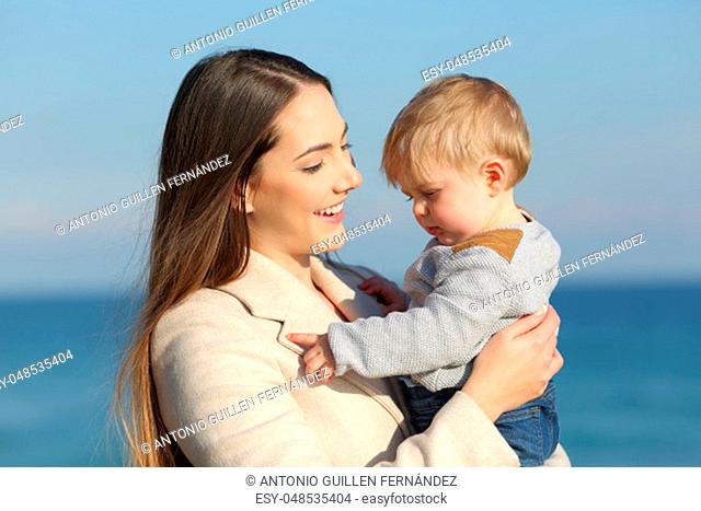 Portrait of a happy mother holding her angry son kid on the beach in a sunny day of winter