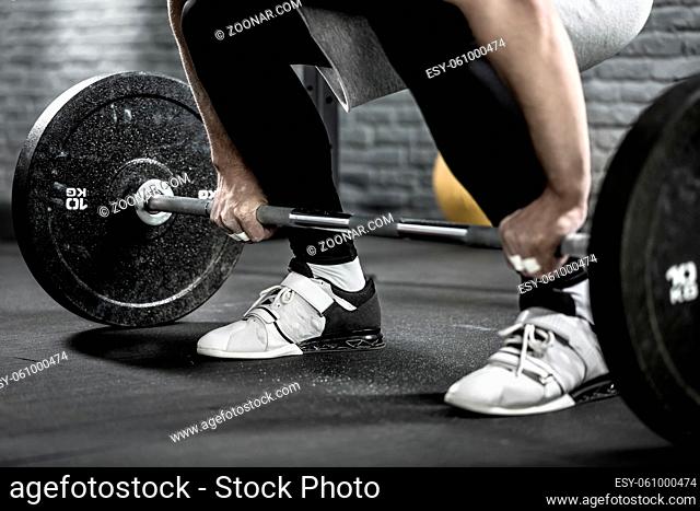 Close-up photo of a barbell and the legs of a man who prepares to raise it from the floor in the gym. He wears sportswear with white sneakers