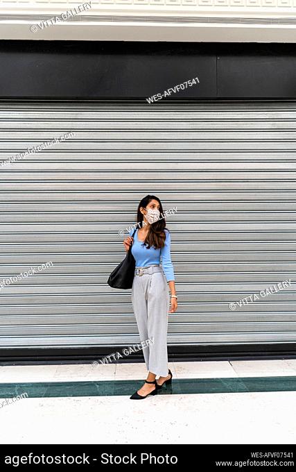 Contemplating woman with purse standing against shutter during COVID-19