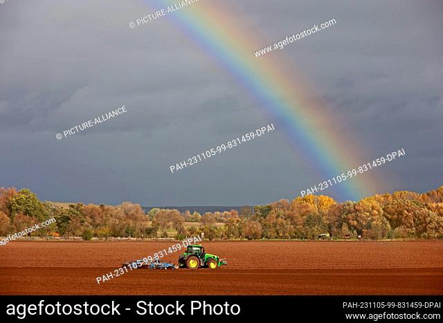 05 November 2023, Saxony-Anhalt, Derenburg: A farmer drives his tractor across a field - a rainbow glows in the background