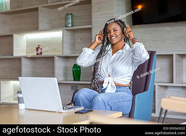 African American woman wearing and holding headphones in home with a laptop in front of her looking ar camera. Concept of people in home