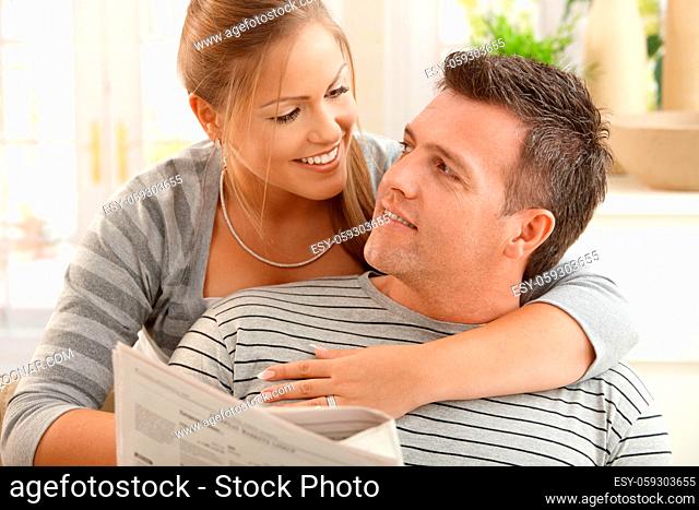 Happy couple sitting at home looking at each other, man holding newspaper, smiling