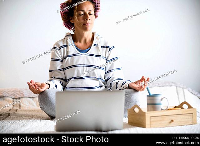 Woman with hair curlers meditating on bed in front of laptop