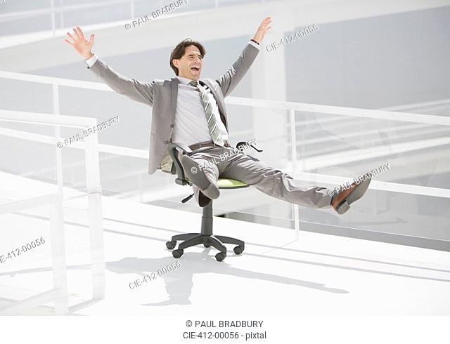Carefree businessman sliding down walkway on office chair with wheels