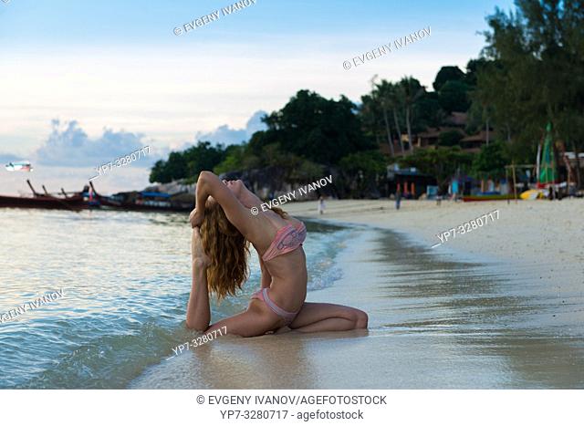Girl practicing yoga in pigeon pose on the beach with Thai longtail boats, Ko Lipe, Thailand