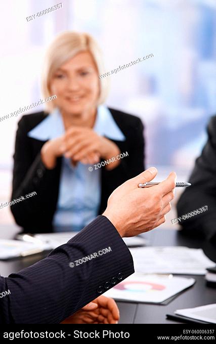Hand in closeup holding pen at business meeting, partner listening in the background