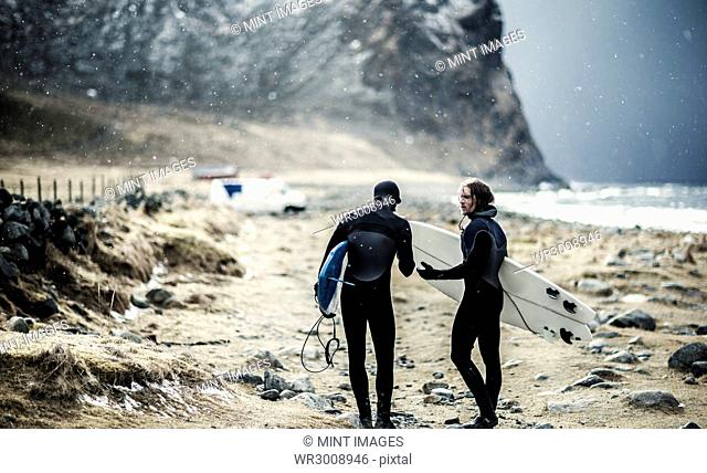 Two surfers wearing wetsuits and carrying surfboards walking towards a van