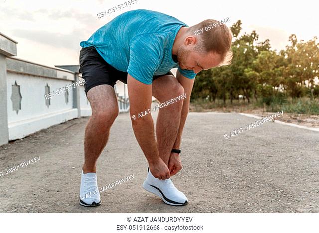 portrait of Caucasian guy in a blue t-shirt and black shorts, who ties his laces on white sports shoes before Jogging
