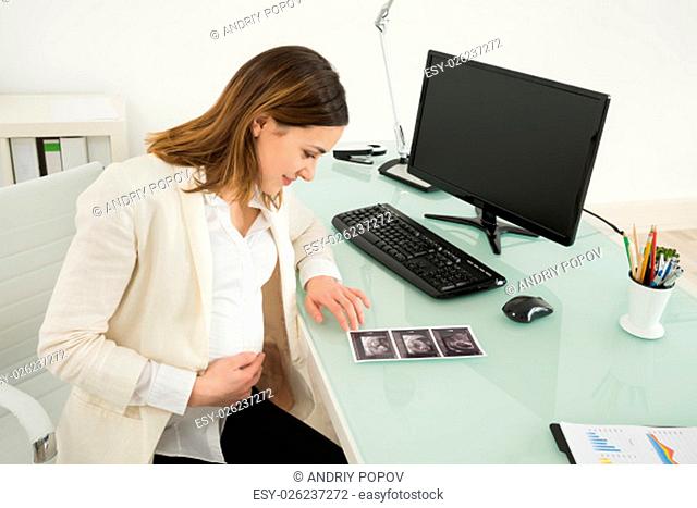Happy Pregnant Businesswoman Looking At Ultrasound Scan Report On Desk