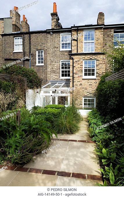 York stone paved path, interspaced with red brick, with clipped box, ornamental grasses, acanthus and alchemilla mollis. Rear view of house and garden in...