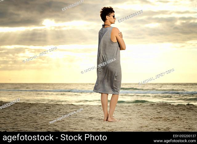 beautiful woman in a gray dress is looking at the sea on the beach at sunset