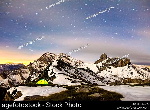 Illuminated camping tent with star trails at night. High altitude alpine landscape with snow covered mountains. Alps, Switzerland