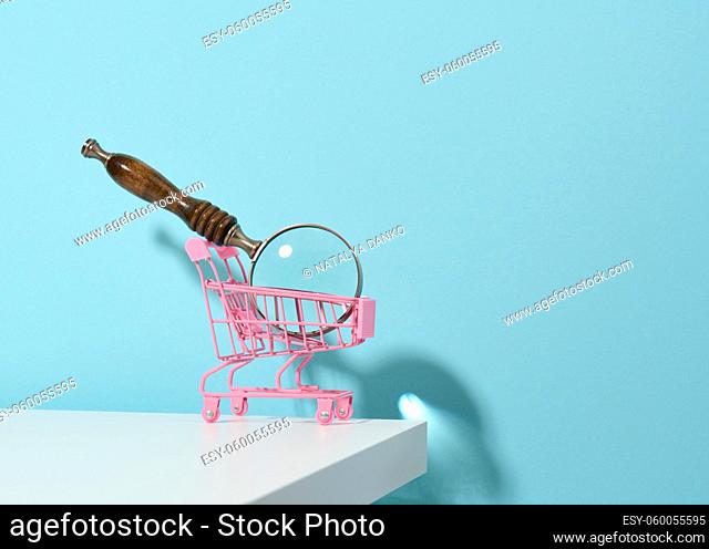 miniature metal pink trolley and wooden magnifier on a light blue background. The concept of search and selection of purchases, savings