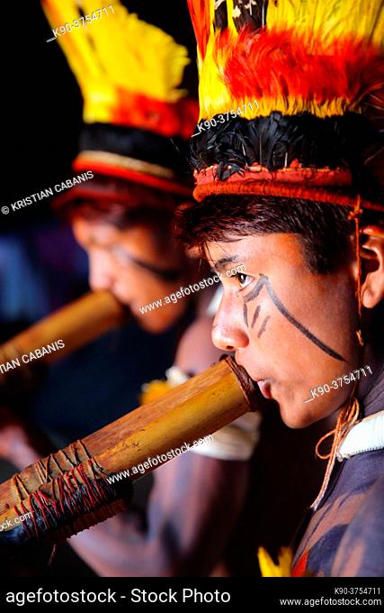 Two native indios in festive clothing and feathres in their hair playing their long pipe inside a hut, Mato Grosso, Brazil, South America