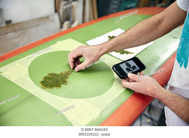 Man creating a design on surfboard laying an oak leaf on the board, using mobile phone in workshop