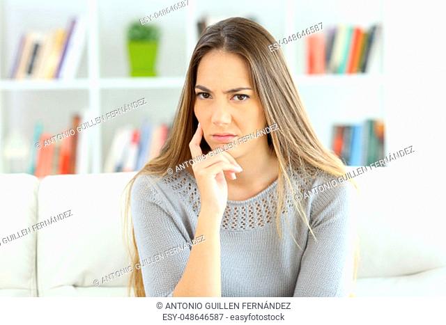 Front view portrait of a suspicious woman looking at camera sitting on a sofa at home