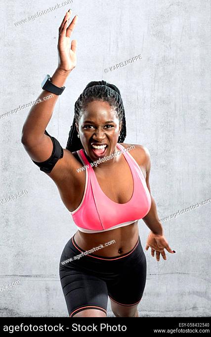 Medium shot of black female athlete in take off position. Young woman with vigorous face expression with hands in air against grey concrete background