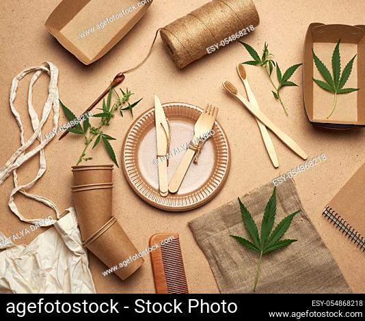 textile bag and disposable tableware from brown craft paper, green hemp leaves on a wooden background. View from above, plastic rejection concept, zero waste