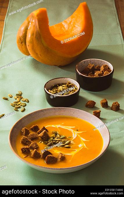 White ceramic bowl of pumpkin cream soup, served with homemade croutons. Piece of orange pumpkin and two bowls with pumpkin seeds and croutons placed behind