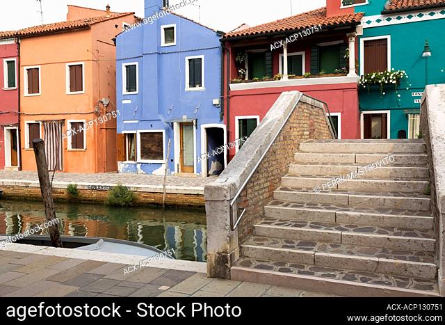Stone and brick footbridge over canal lined with colourful stucco houses decorated with striped curtains over entrance doors and flowers, Burano Island
