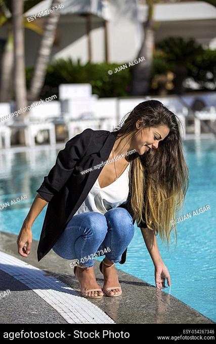 Elegant executive girl in jacket relaxing on hotel territory and touching water in refreshing pool and smiling