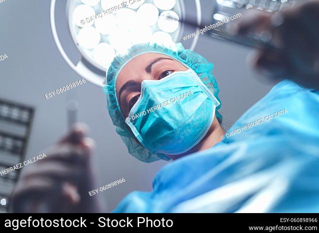 From below woman surgeon in medical uniform using professional tools while standing under bright light in operating theater. High quality photo