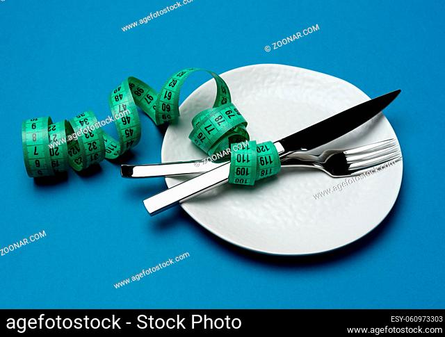 white round plate and fork and knife wrapped in green measuring tape on blue background, weight loss concept, flat lay