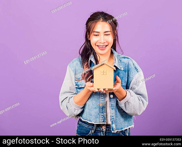 Happy Asian portrait beautiful cute young woman wear denim excited smiling holding house model on hand, studio shot isolated on purple background