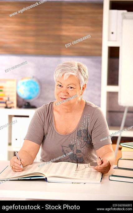 Portrait of senior teacher sitting at desk in classroom, searching in a book, looking at camera