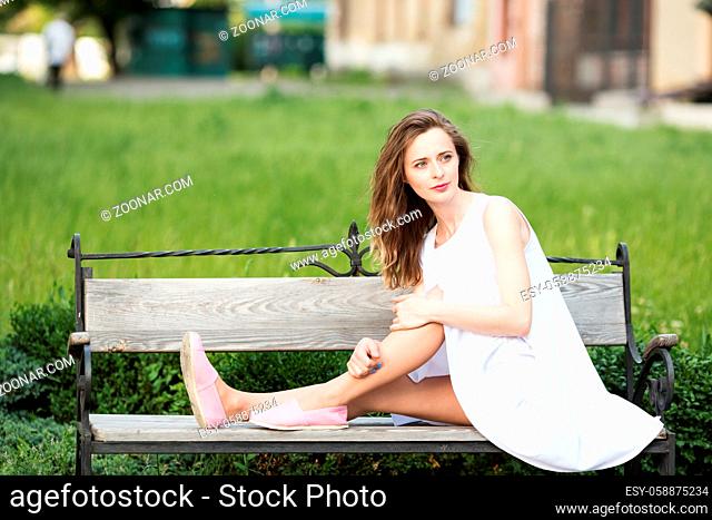 Lovely urban girl in short white dress in the street. Portrait of a happy smiling woman. Fashionable blonde girl sitting on a bench in a city park