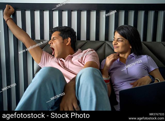 A young newlywed couple in bedroom with the husband yawning and wife using laptop